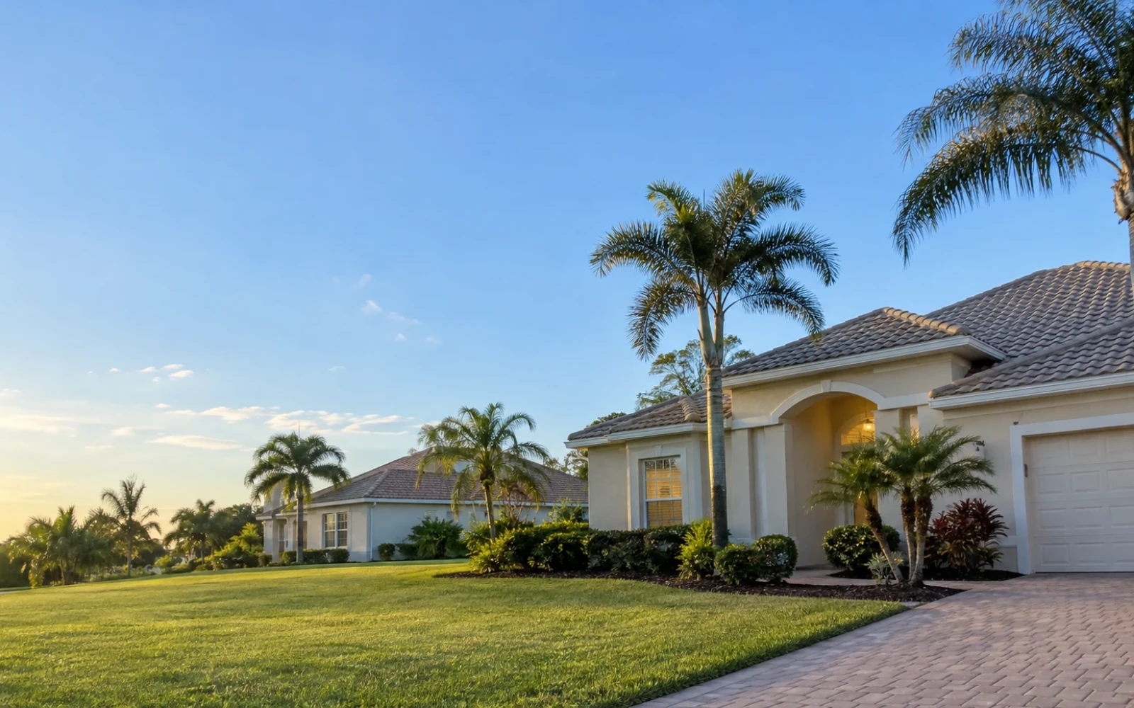 An ordinary residential street in Southwest Florida with two single-storey homes, palm trees and trimmed lawns under a clear blue sky, with no vehicles.