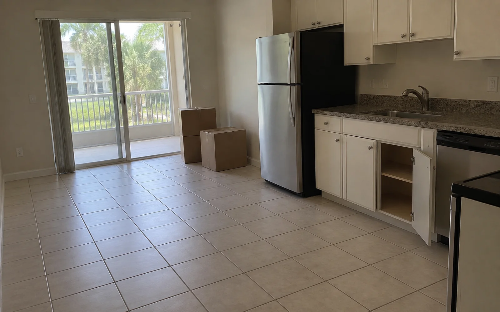An empty condominium living area and kitchen with tiled floors, stacked cardboard boxes, a stainless steel refrigerator, an open lower cabinet, and sliding doors to a balcony.
